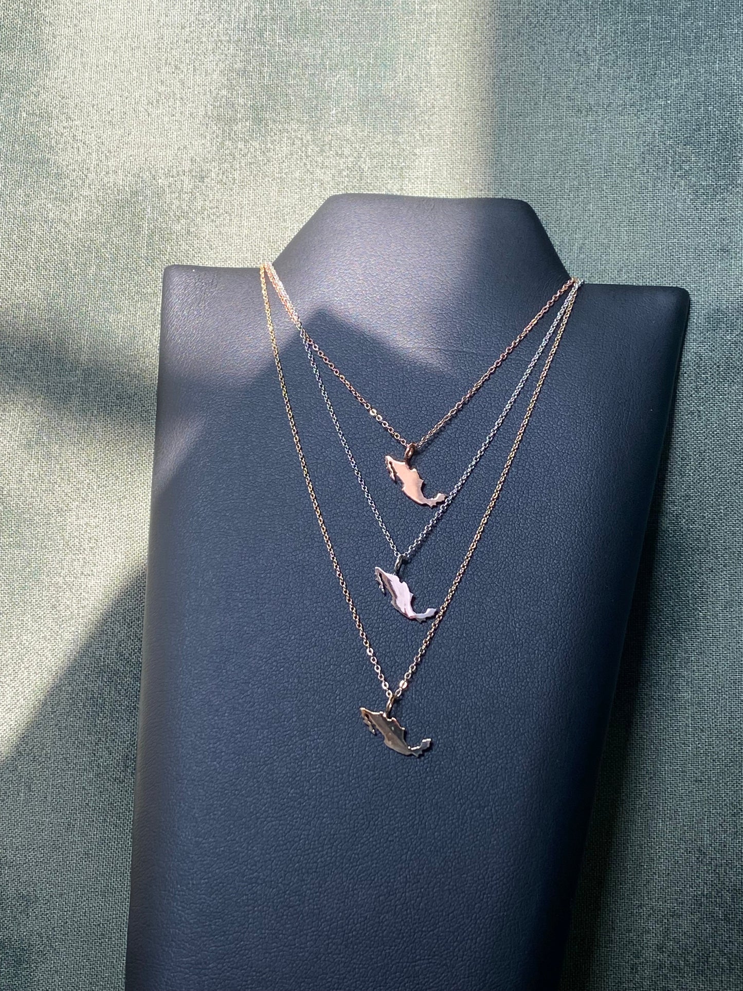 Display of layered Mexico map pendant necklaces in gold, rose gold, and silver on black jewelry bust, showcasing delicate chain styles.