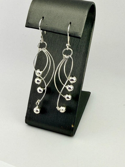 Sterling silver dangle earrings with curved wire design and polished silver bead accents displayed on a black jewelry stand.